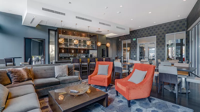 Modern lounge with a grey sectional, two orange armchairs, coffee table, and a bar. Blue patterned rug and walls.