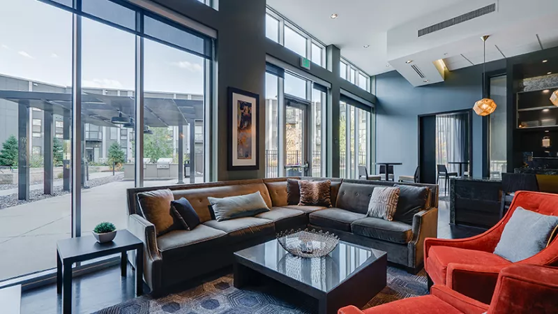 Spacious, modern lounge with large windows, an L-shaped sofa, coffee table, and two vibrant orange armchairs. Overlooks a patio.