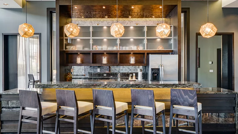 Modern bar with granite counter, six wooden stools, and warm spherical pendant lights over shelves with glassware and appliances.