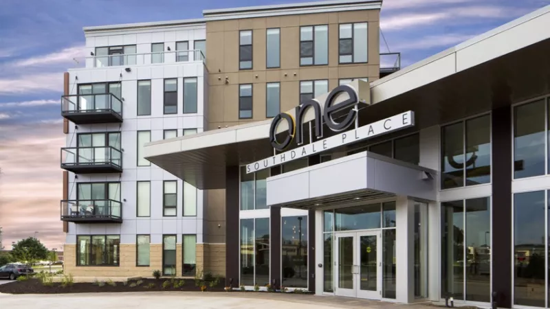 Modern multi-story building with white and brown facades, balconies, and a glass entrance with "one" above, under a cloudy evening sky.