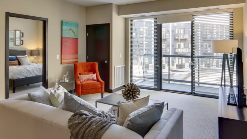 Modern living room with a white sofa, orange accent chair, abstract art, and sliding glass doors leading to a balcony.