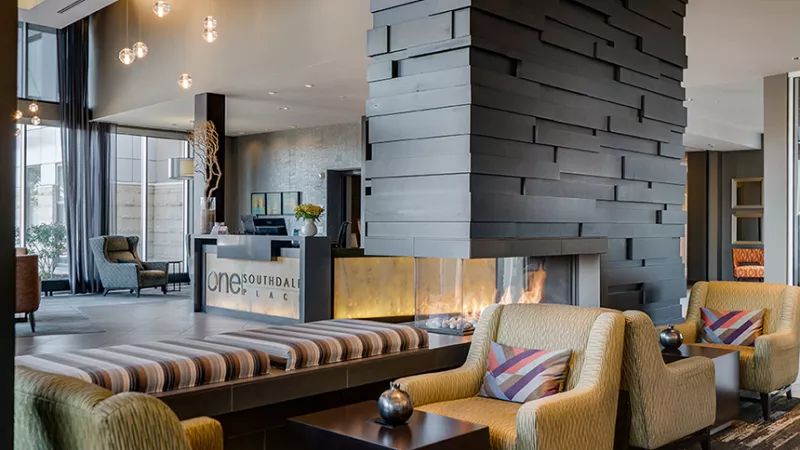 Contemporary lobby with a tall, dark, textured fireplace, inviting armchairs, and a bright reception desk in the background.
