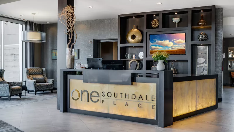 Modern lobby reception desk, displaying "ONE SOUTHDALE PLACE" logo, with decorative shelving and a seating area.