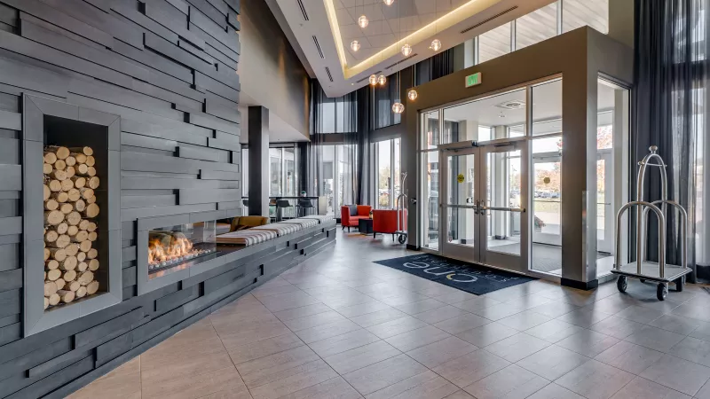 Modern hotel lobby with a contemporary grey panel wall, a fireplace, stacked logs, and a wide glass entrance.