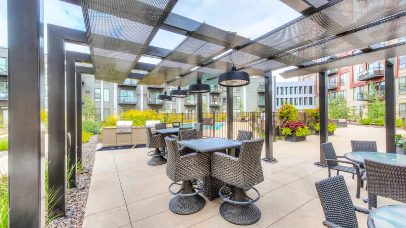 Outdoor modern patio with a pergola, tables, chairs, and a grill, surrounded by greenery and apartment buildings.