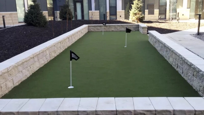Outdoor putting green with three black flags, stone walls, and modern apartment buildings in the background.