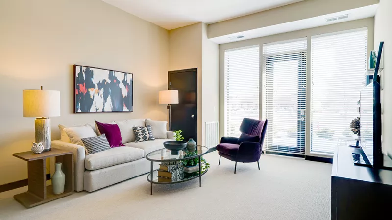 Bright, modern living room with cream walls, white sofa, purple armchair, glass coffee table, and large sunlit windows.