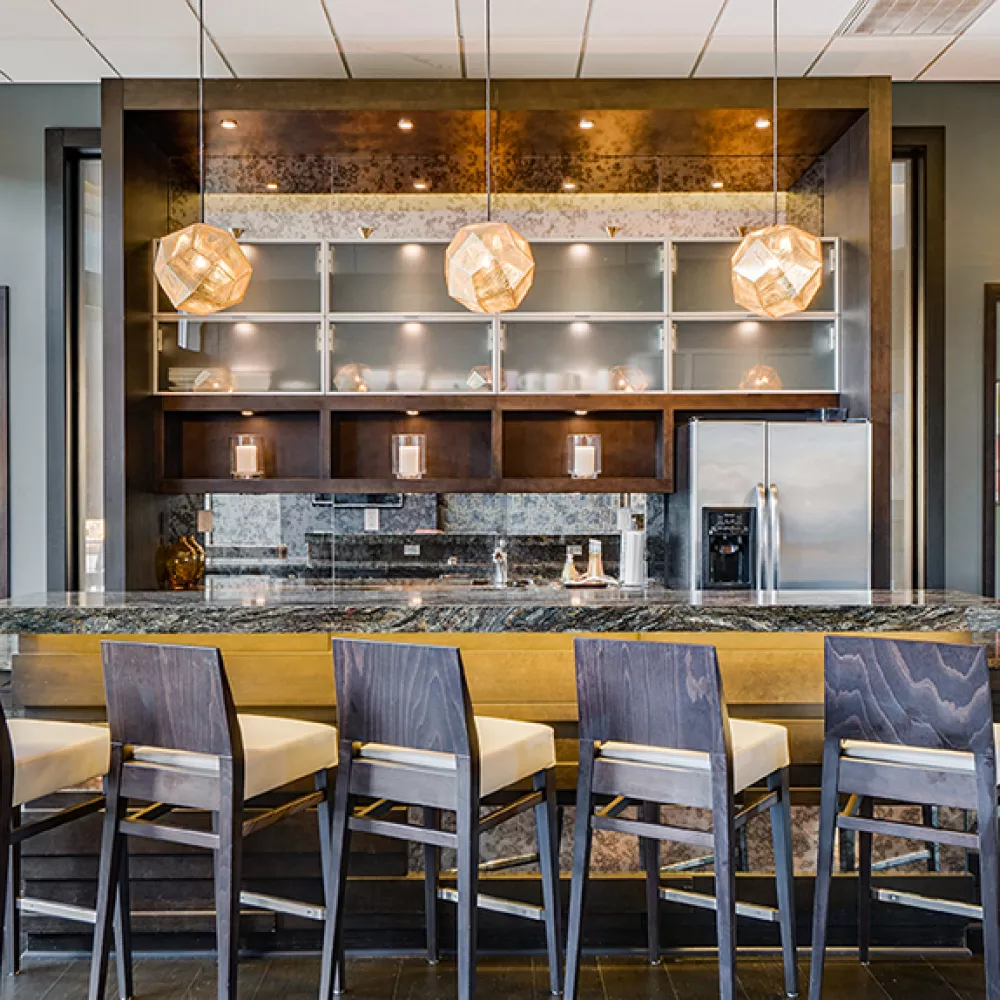 Modern bar with granite counter, six wooden stools, and warm spherical pendant lights over shelves with glassware and appliances.