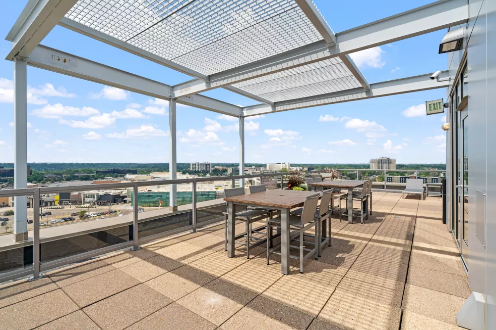 Bright rooftop patio with two dining tables and chairs under a metal pergola, city skyline visible under a blue sky.