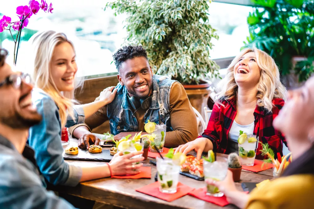 Diverse friends laughing heartily while socializing at a restaurant table with drinks.