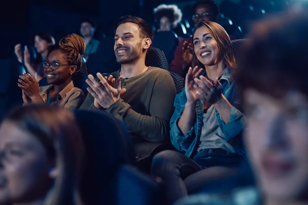 Photograph: Diverse audience claps and smiles in a dark movie theater.