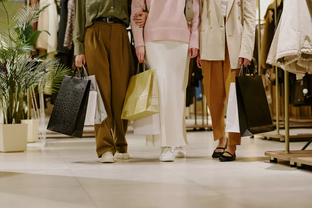 Three shoppers, seen from the waist down, walk together through a bright clothing store, holding several shopping bags.